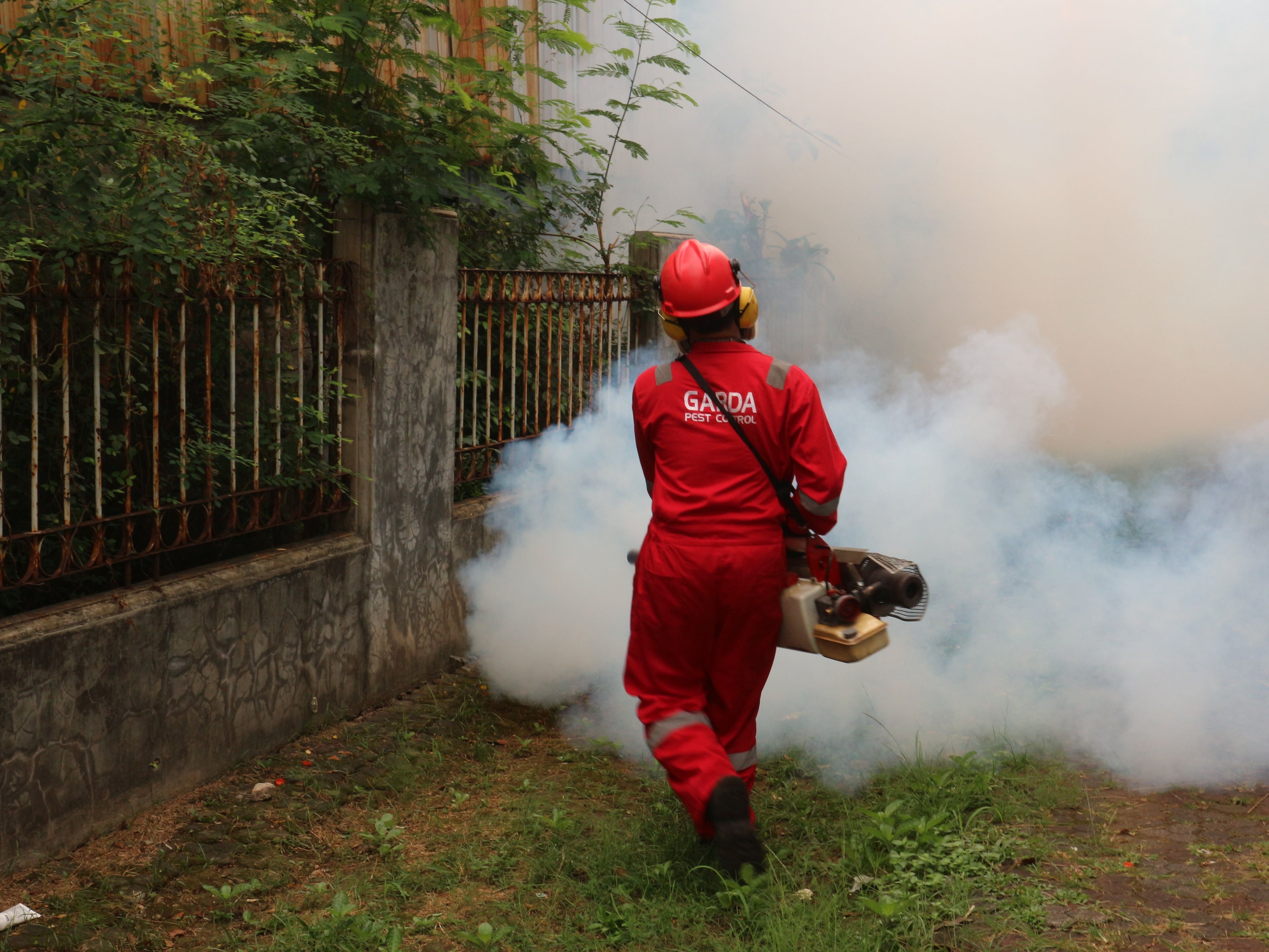 Pest control technician in red coveralls, hard hat, and ear protection fogging the exterior of a residential property