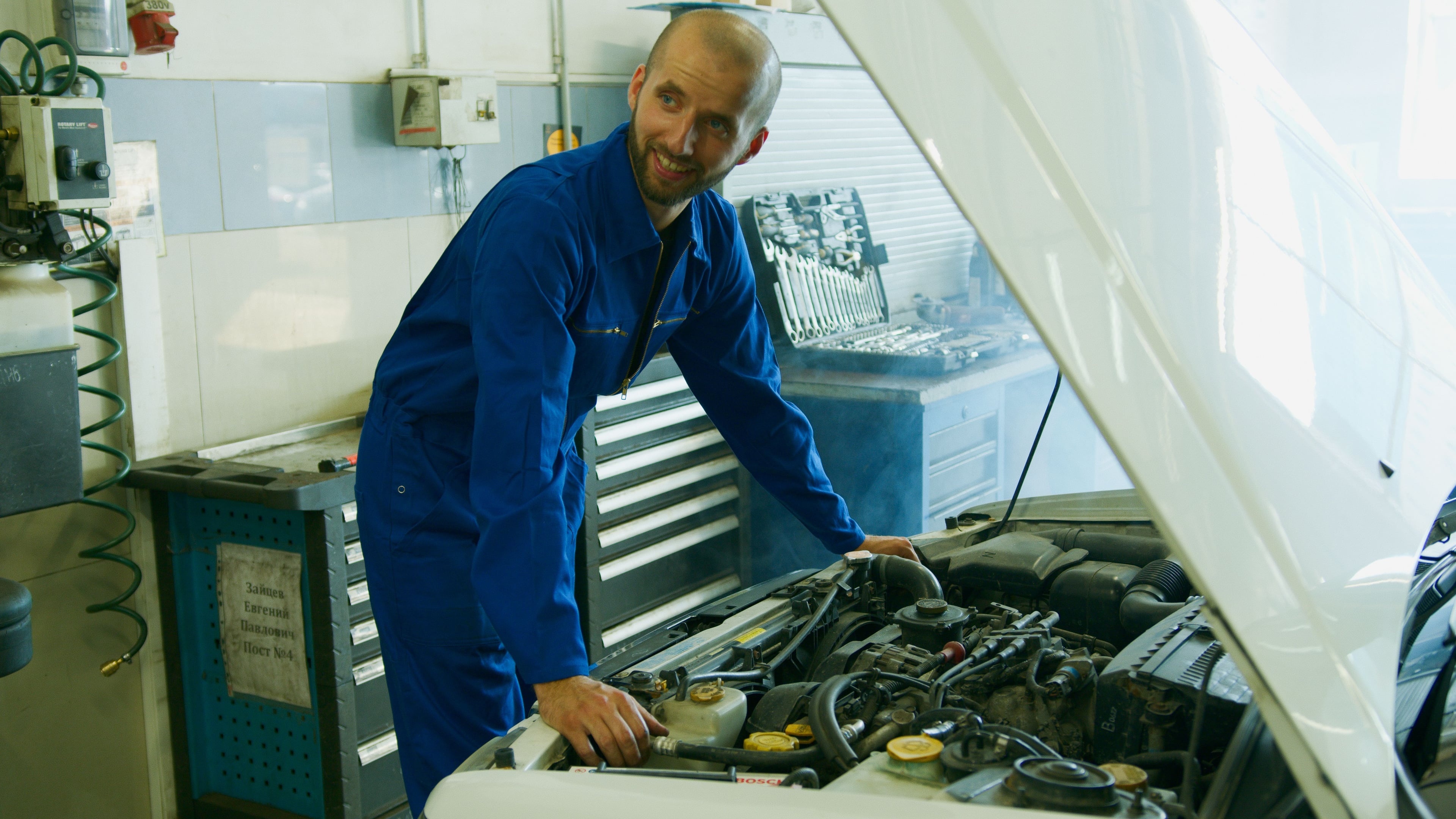 Friendly smiling auto mechanic in blue coveralls working on a car engine under the open hood in a clean repair shop